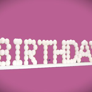 May include: White "BIRTHDAY" sign with a bubble-like font, mounted on a white base. The letters are made of small, round, white shapes. The background is a solid, medium-dark pink.