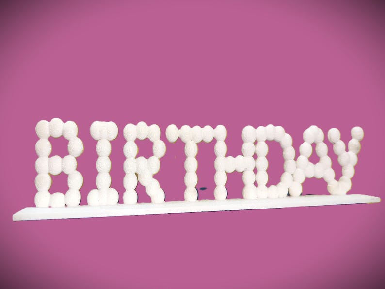 May include: White "BIRTHDAY" sign with a bubble-like font, mounted on a white base. The letters are made of small, round, white shapes. The background is a solid, medium-dark pink.