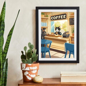 May include: Framed art print featuring a whimsical illustration of a black and white cat working in a coffee shop. The print includes a "COFFEE" sign, blue chairs, and a counter with pastries. The artwork is displayed on a wooden table with a cactus and other decorative items.