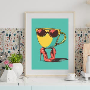 May include: Framed art print featuring a yellow coffee cup wearing sunglasses and red high heels. The cup is set against a teal background. The artwork has a retro, pop art style, with bold colors and clean lines. The frame is a light wood color.
