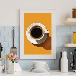 May include: A framed art print featuring a top-down view of a white coffee cup and saucer filled with dark coffee. The background is a solid orange color. The print is displayed in a kitchen setting with other kitchen items.