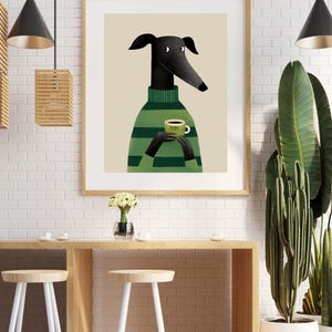May include: A framed art print featuring a stylized greyhound dog wearing a green striped sweater and holding a green coffee cup. The artwork has a beige background and is displayed in a light wood frame. The cup has the word "TUMI" on it.
