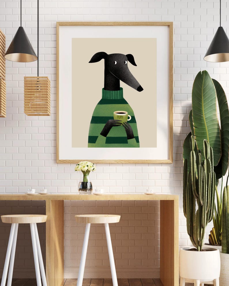 May include: A framed art print featuring a stylized greyhound dog wearing a green striped sweater and holding a green coffee cup. The artwork has a beige background and is displayed in a light wood frame. The cup has the word "TUMI" on it.