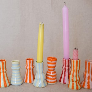 Op de afbeelding: Een collectie van elf keramische kandelaars met diverse handgeschilderde streeppatronen in levendige kleuren zoals oranje, rood, lichtblauw en geel. Drie van de houders bevatten taps toelopende kaarsen in geel, roze en lichtroze.
