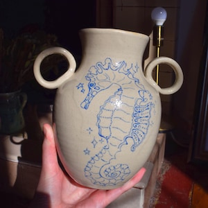 May include: A ceramic vase with two handles, painted in a light beige color with a blue seahorse design. The seahorse is outlined in blue and has a detailed design with stars and swirls.