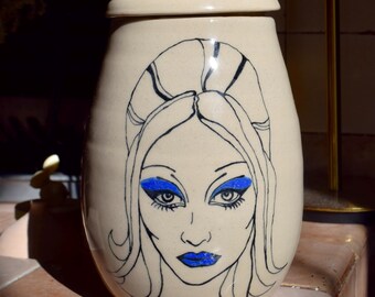 May include: A ceramic jar with a lid, featuring a hand-painted illustration of a woman with blue eyeshadow and blue lipstick. The jar is cream-colored with black outlines.