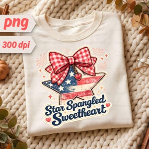 Puede incluir: Una camiseta color crema con un gráfico de un lazo de cuadros vichy rojo y blanco sobre una estrella con la bandera estadounidense. Un pequeño dije de corazón rojo cuelga del lazo. El texto "Star Spangled Sweetheart" aparece en azul.