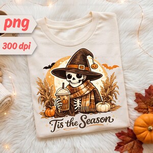 May include: A cream-colored t-shirt featuring a graphic of a skeleton wearing a witch hat and a plaid scarf, holding a coffee cup. The design includes corn stalks, pumpkins, bats, and a full moon, with the text 'Tis the Season' printed below.
