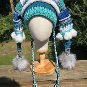 May include: A crochet jester hat featuring two long, pointed tails in shades of teal, blue, white, and grey. The hat is accented with white faux fur trim, small white pom-poms, and large grey faux fur pom-poms at the ends of the braided tassels.