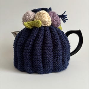 May include: A navy blue knitted teapot cozy with a flower design. The flowers are made of yarn in shades of pink, cream, and purple.