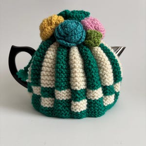 Puede incluir: Un calienta-tetera de punto de rayas verdes y blancas con un diseño floral en la parte superior. Las flores son amarillas, verde azulado, rosas y verdes.