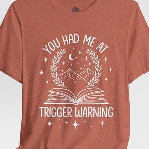 Può includere: T-shirt color ruggine con grafica bianca. La grafica presenta la scritta "YOU HAD ME AT TRIGGER WARNING" sopra un libro aperto, montagne e stelle. È inclusa anche una luna crescente.