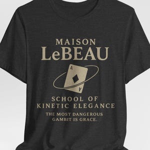May include: Dark gray t-shirt with the text "MAISON LeBEAU" above a playing card design and the words "SCHOOL OF KINETIC ELEGANCE". The phrase "THE MOST DANGEROUS GAMBIT IS GRACE" is printed below the design.