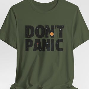 以下が含まれることがあります： オリーブグリーンのTシャツ。黒いブロック体で「DON'T PANIC」と書かれています。文字は星空のデザインで、黄色い太陽や他の天体で埋められています。