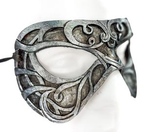 Può includere: Una maschera da ballo argentata con intricati disegni floreali. La maschera ha un'unica apertura per l'occhio ed è realizzata in un materiale leggero.
