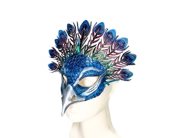 May include: A peacock-inspired mask featuring a textured blue scale pattern across the face, a silver beak, and a prominent fan of iridescent blue, green, and purple feathers extending from the top. The mask includes eye cutouts and a blue tie.