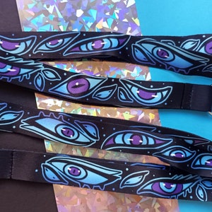 May include: Two black lanyards with silver clasps. The lanyards are printed with a repeating pattern of blue and purple eyes.