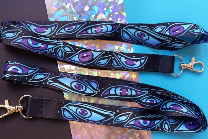 May include: Two black lanyards with silver clasps. The lanyards are printed with a repeating pattern of blue and purple eyes.