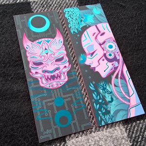 Può includere: Due segnalibri illustrati. Uno presenta un design di maschera Oni rosa e blu, l'altro una donna cyborg rosa e blu. Entrambi hanno uno sfondo grigio con accenti turchese e un motivo a circuito stampato.
