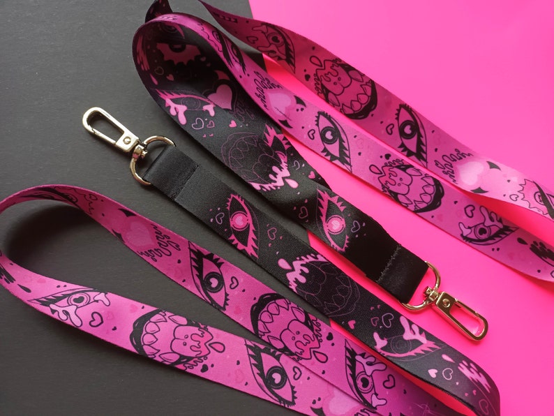 May include: Two pink and black patterned lanyards with a gold metal clasp. One lanyard has a black background with pink eyes and dripping hearts. The other lanyard has a pink background with black eyes and dripping hearts.