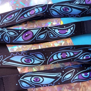 May include: A black lanyard with a repeating pattern of blue and purple eyes with white highlights. The eyes are stylized and have a whimsical feel. The lanyard has a silver metal clasp.