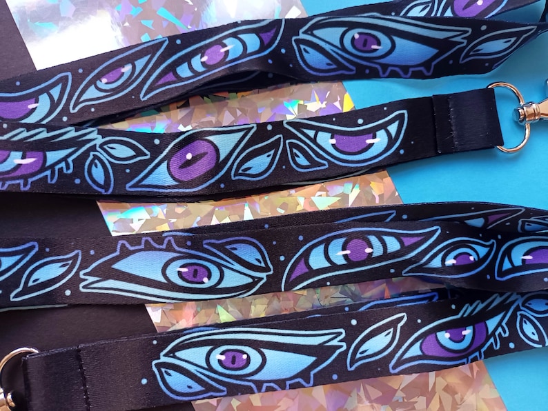 May include: A black lanyard with a repeating pattern of blue and purple eyes with white highlights. The eyes are stylized and have a whimsical feel. The lanyard has a silver metal clasp.
