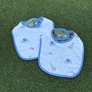 May include: Two white baby bibs with blue trim. The bibs feature a pattern of elephants, ladybugs, and dinosaurs. The bibs are made of soft, absorbent fabric.