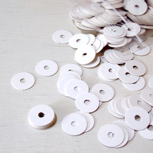 May include: A large collection of small, flat, circular white paper discs, each featuring a single centered hole. These round white paper cutouts are scattered across the surface, spilling out from a clear plastic bag at the top of the frame.