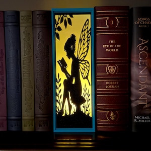 May include: A rectangular light box with a bright yellow background featuring a black silhouette of a winged fairy reading a book. The light box is framed in blue and positioned between several hardcover books on a shelf.