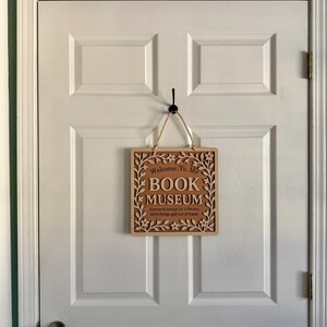 Wooden "Book Museum" sign hanging on a white paneled door by twine, shown in a real home setting with soft natural lighting.