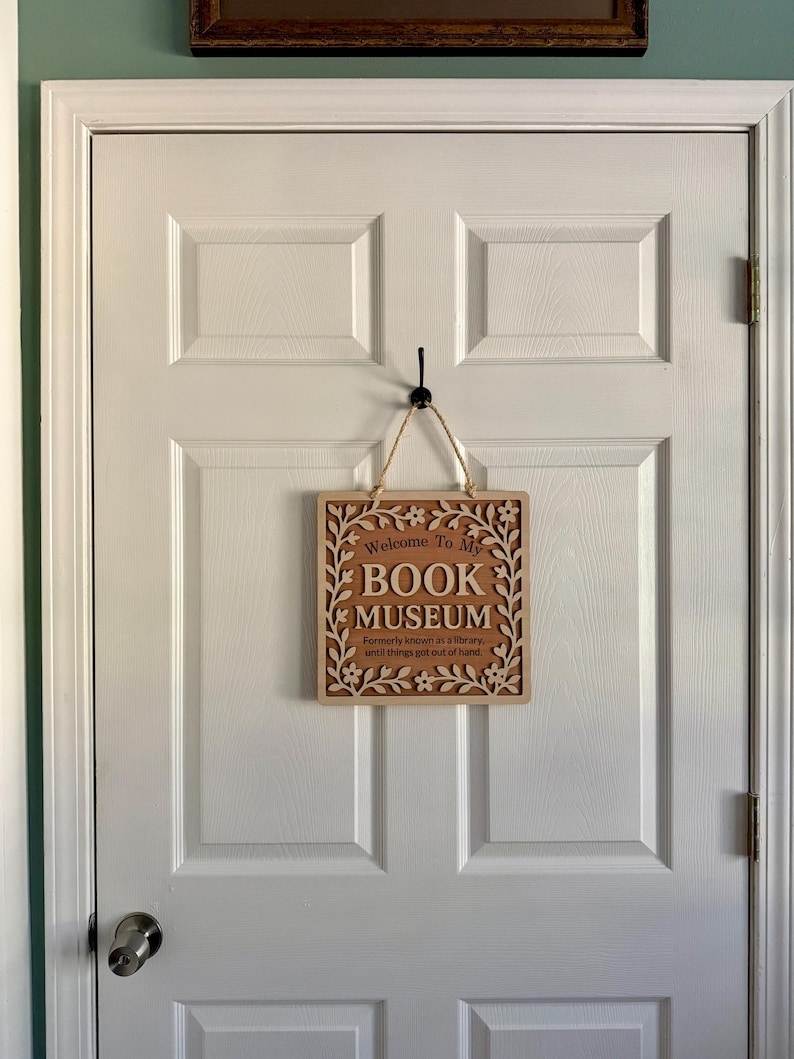 Wooden "Book Museum" sign hanging on a white paneled door by twine, shown in a real home setting with soft natural lighting.