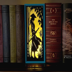 May include: A rectangular light box with a bright yellow glow featuring a black silhouette of a figure wearing a conical hat and holding a sword amidst bamboo stalks. The light box is framed in blue and positioned between several hardcover books.