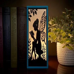 May include: A rectangular light box with a teal frame featuring a black silhouette of a fairy with wings sitting and reading a book. The internal light illuminates the scene, highlighting the intricate cutout details of the fairy and foliage.