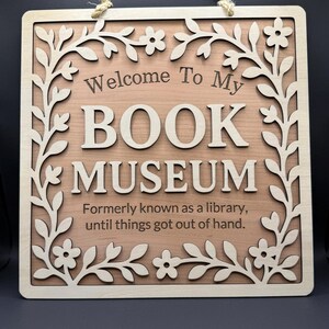 Close-up of a wooden sign that says "Welcome to My Book Museum," with a layered floral border and engraved lettering, displayed against a black backdrop.