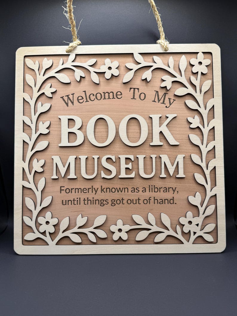 Close-up of a wooden sign that says "Welcome to My Book Museum," with a layered floral border and engraved lettering, displayed against a black backdrop.