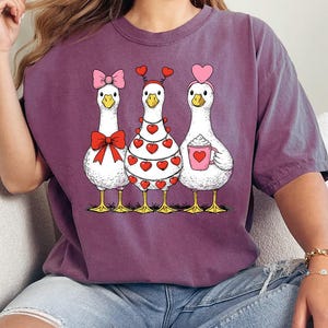 May include: A purple t-shirt featuring three cartoon geese with Valentine's Day themed decorations. One goose has a pink bow, another has a heart-covered dress, and the third holds a mug. The shirt has the text "Flower Crafts StoreUS".