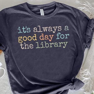 May include: A dark charcoal gray short-sleeve t-shirt featuring the text "it's always a good day for the library" printed in a multi-colored, retro-inspired font with shades of light blue, yellow, orange, and white lettering on the chest.