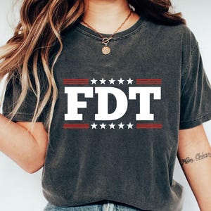 May include: Dark gray t-shirt with the white letters "FDT" in a bold font, framed by red stripes and white stars. The shirt is a casual style, suitable for everyday wear.