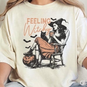 May include: A cream-colored t-shirt featuring a graphic illustration of a person in a witch hat sitting in a wooden chair, holding a cup, with a black cat, bats, a jack-o'-lantern, and the text "FEELING Witchy" printed in black and orange.