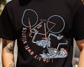 以下が含まれることがあります： 自転車から落ちる骸骨のグラフィックが描かれた黒いTシャツ。骸骨は青いショートパンツと赤いキャップを着用している。自転車と骸骨のイラストの周りには、ピンク色の曲線的なフォントで「i do my own stunts」という文字がプリントされている。
