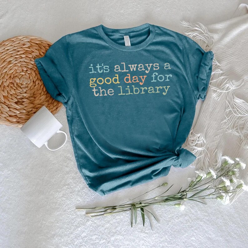以下が含まれることがあります： クルーネックの青緑色の半袖Tシャツ。Tシャツの中央には、「it's always a good day for the library」というテキストが、白、黄色、オレンジ、ライトグリーンの色合いのマルチカラーフォントでプリントされています。