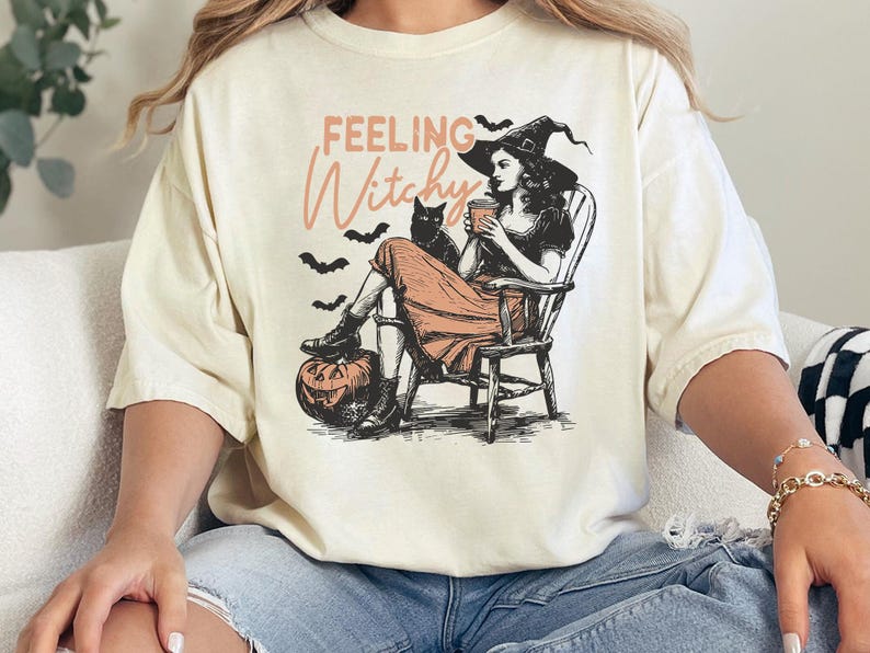 May include: A cream-colored t-shirt featuring a graphic illustration of a person in a witch hat sitting in a wooden chair, holding a cup, with a black cat, bats, a jack-o'-lantern, and the text "FEELING Witchy" printed in black and orange.