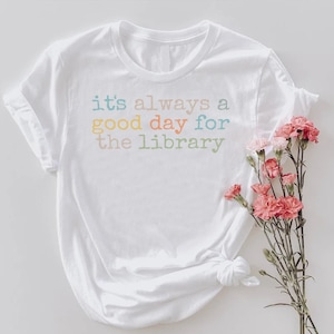 以下が含まれることがあります： 胸元に「it's always a good day for the library」とマルチカラーの文字がプリントされた白い半袖Tシャツ。Tシャツの横には、緑の茎と葉がついたピンクのカーネーションの小さな花束が添えられています。
