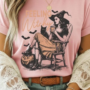 May include: A light pink t-shirt featuring a black and peach graphic illustration of a person in a witch hat sitting in a wooden chair, holding a cup, with a black cat, bats, and a jack-o'-lantern. The text "FEELING Witchy" is printed above.