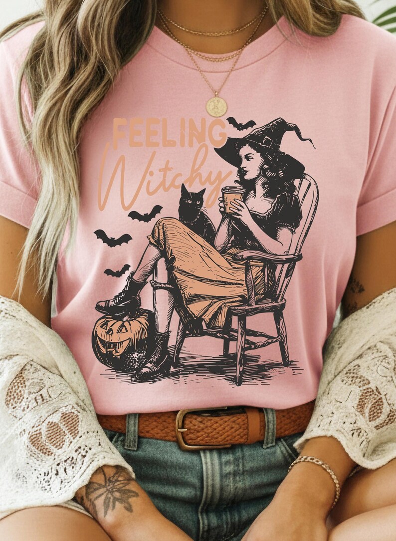 May include: A light pink t-shirt featuring a black and peach graphic illustration of a person in a witch hat sitting in a wooden chair, holding a cup, with a black cat, bats, and a jack-o'-lantern. The text "FEELING Witchy" is printed above.