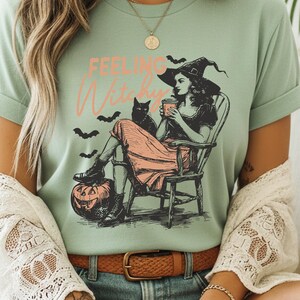 May include: A sage green t-shirt featuring a graphic print of a person in a witch hat sitting in a wooden chair, holding a cup, with a black cat, bats, and a jack-o'-lantern. The text "FEELING Witchy" is printed in orange and pink above the illustration.