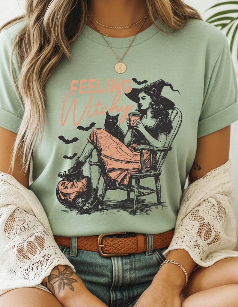 May include: A sage green t-shirt featuring a graphic print of a person in a witch hat sitting in a wooden chair, holding a cup, with a black cat, bats, and a jack-o'-lantern. The text "FEELING Witchy" is printed in orange and pink above the illustration.