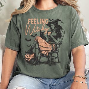 May include: An olive green short-sleeve t-shirt featuring a graphic print of a person in a witch hat sitting in a rocking chair with a black cat, a pumpkin, and bats. The text "FEELING Witchy" is printed in a light orange script and block font.