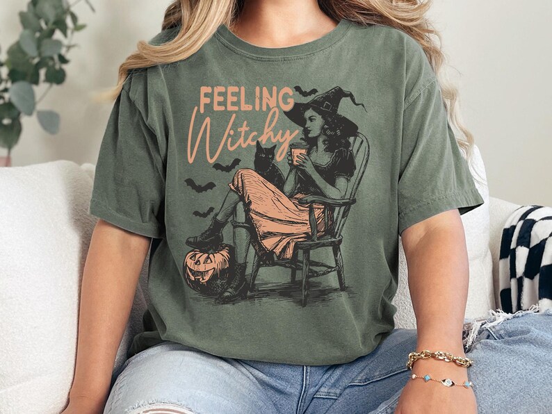 May include: An olive green short-sleeve t-shirt featuring a graphic print of a person in a witch hat sitting in a rocking chair with a black cat, a pumpkin, and bats. The text "FEELING Witchy" is printed in a light orange script and block font.