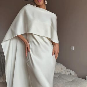 May include: A white satin gown with a cape overlay. The dress has a simple, elegant design with a floor-length skirt. The cape drapes over the shoulders, creating a sophisticated silhouette. The model is wearing pearl earrings.